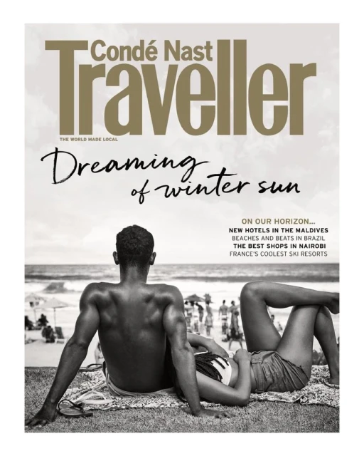 Condé Nast Traveller cover: a back view of a man and a woman lying on the beach, with the ocean and other beachgoers behind them.