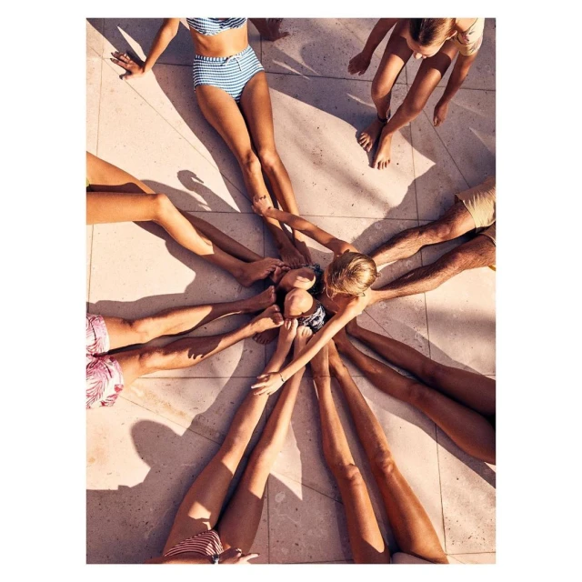 Overhead shot of group of people in swimwear with outstretched legs touching in the center on a tiled surface in bright sunlight.