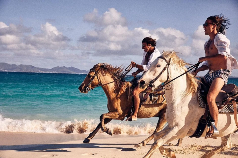Two shirtless men in swim trunks ride horses on a sandy beach along the ocean with blue water and a cloudy sky in a sunny landscape.
