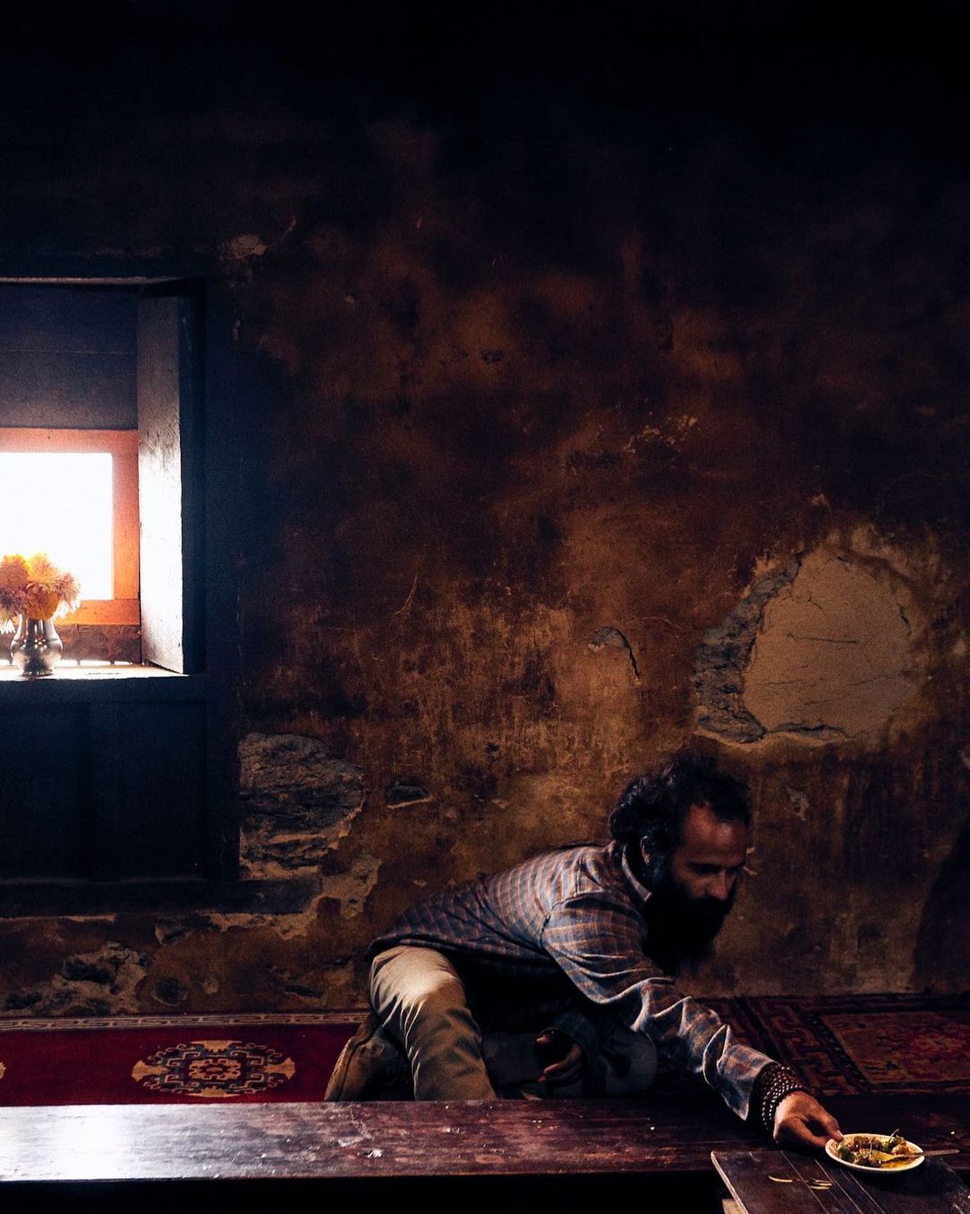 An adult male with a beard reaches for a plate on a dark wooden table in an interior room with a window and textured walls.