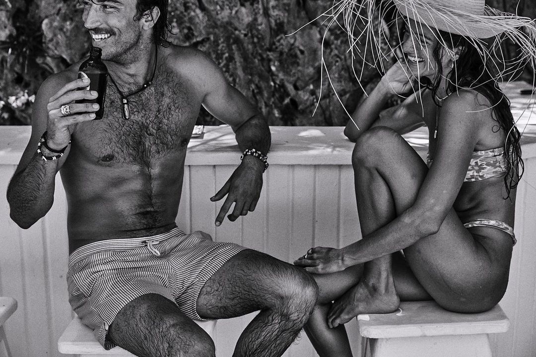 Black-and-white photo of a smiling man holding a bottle next to a smiling woman in a straw hat sitting outside.