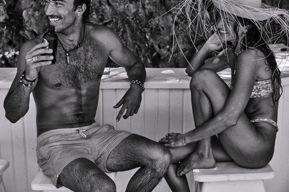 Black-and-white photo of a smiling man holding a bottle next to a smiling woman in a straw hat sitting outside.