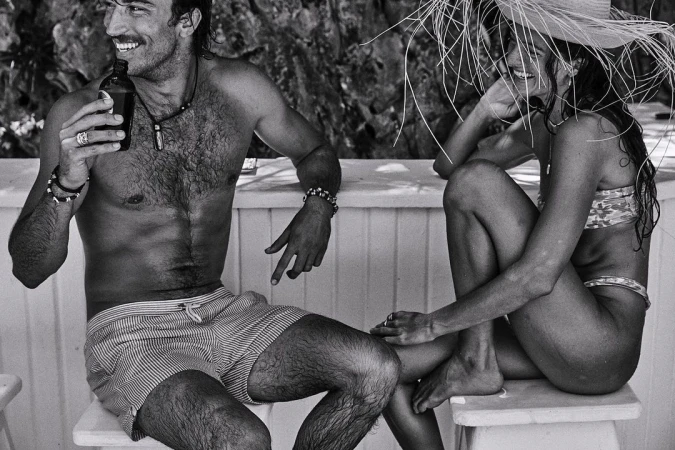 Black-and-white photo of a smiling man holding a bottle next to a smiling woman in a straw hat sitting outside.