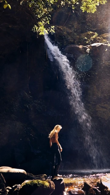 Woman standing near a waterfall in a rocky, forested landscape, capturing the beauty and serenity of nature and adventure.