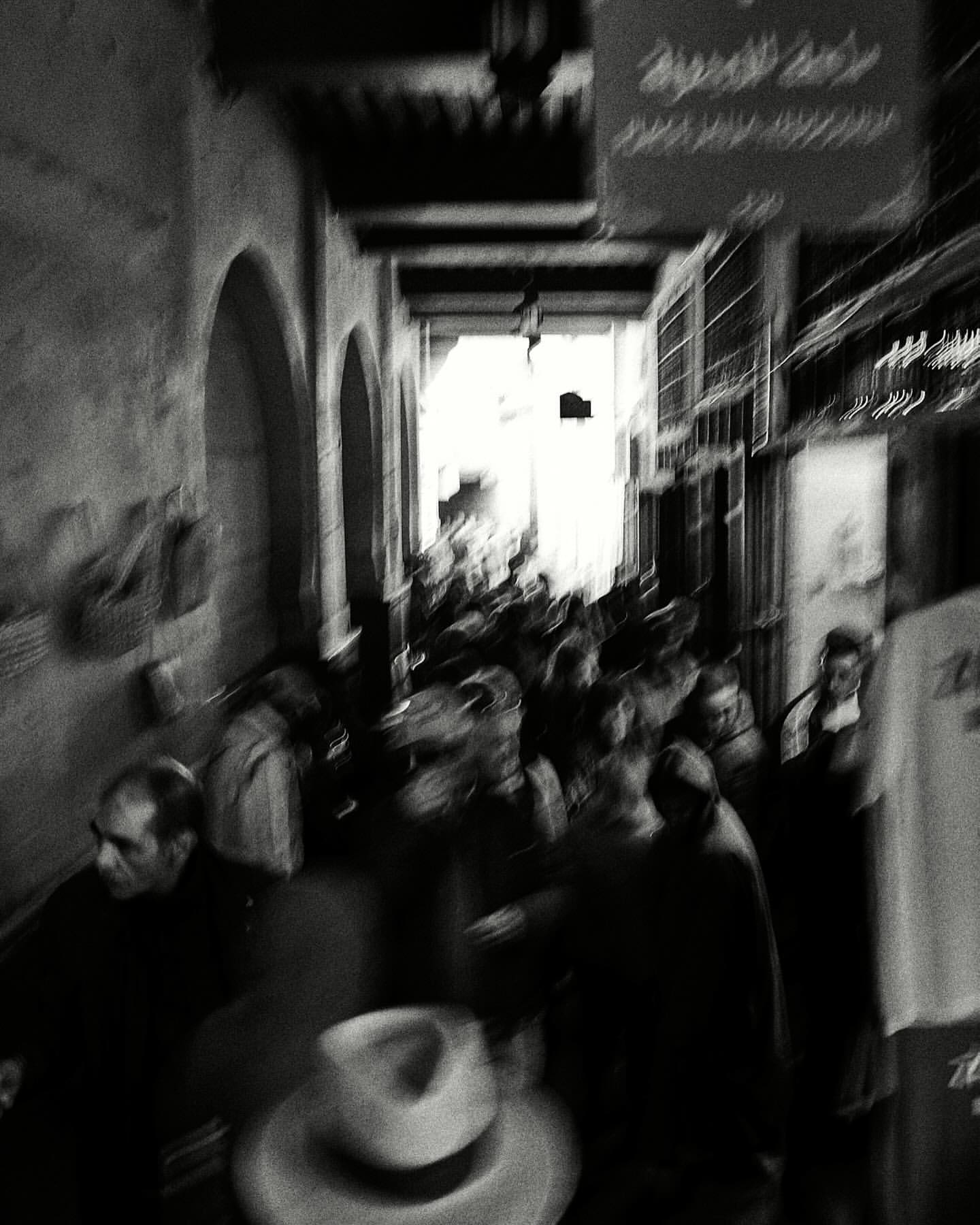 Monochrome photograph of a crowded market street with blurred motion and a hat in the foreground, evoking a sense of movement.