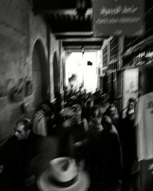 Monochrome photograph of a crowded market street with blurred motion and a hat in the foreground, evoking a sense of movement.
