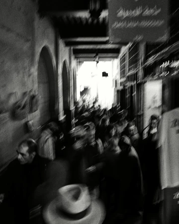 Monochrome photograph of a crowded market street with blurred motion and a hat in the foreground, evoking a sense of movement.