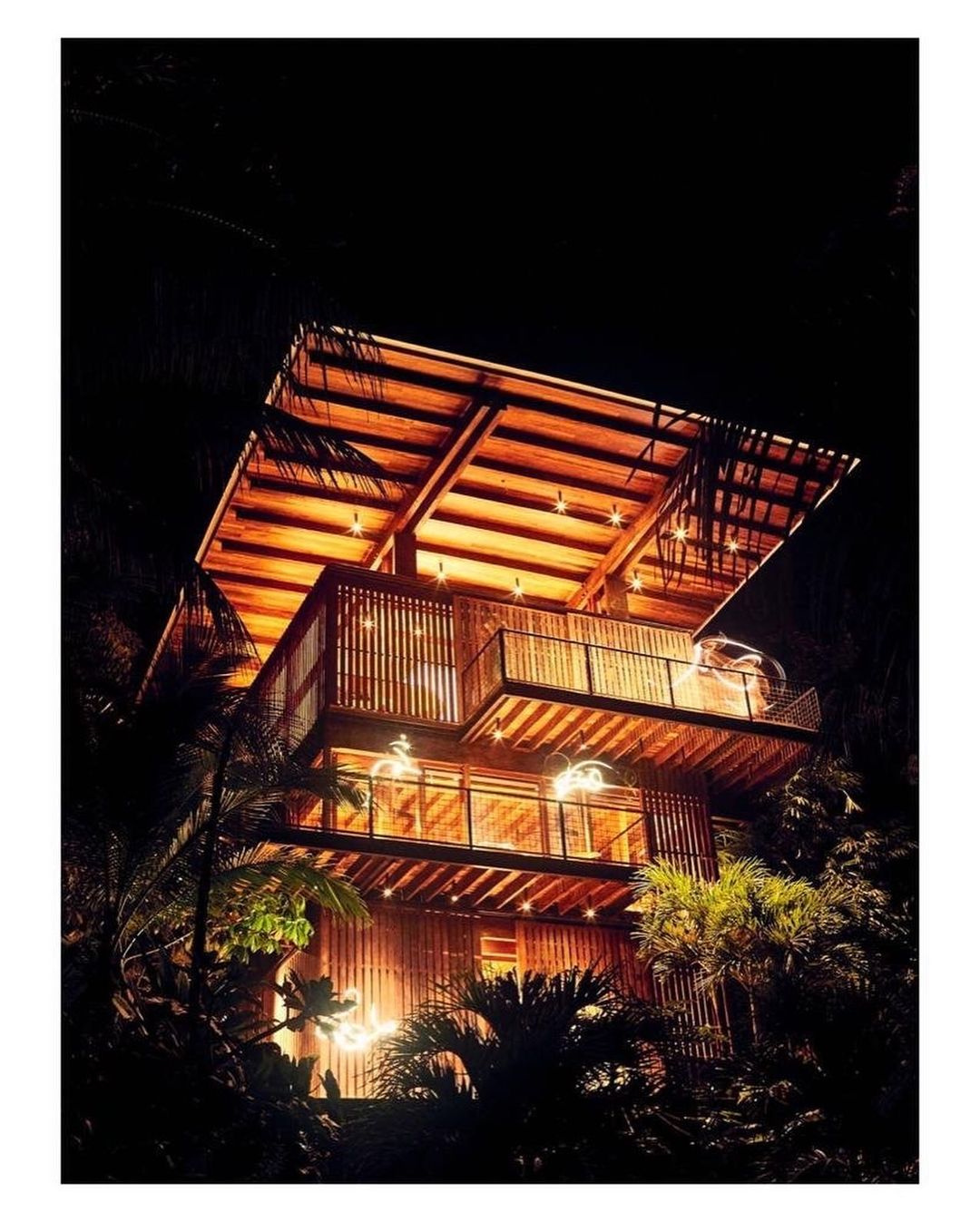 Three-story wooden house with bamboo siding and illuminated balconies stands on a hill at night, surrounded by tropical trees.