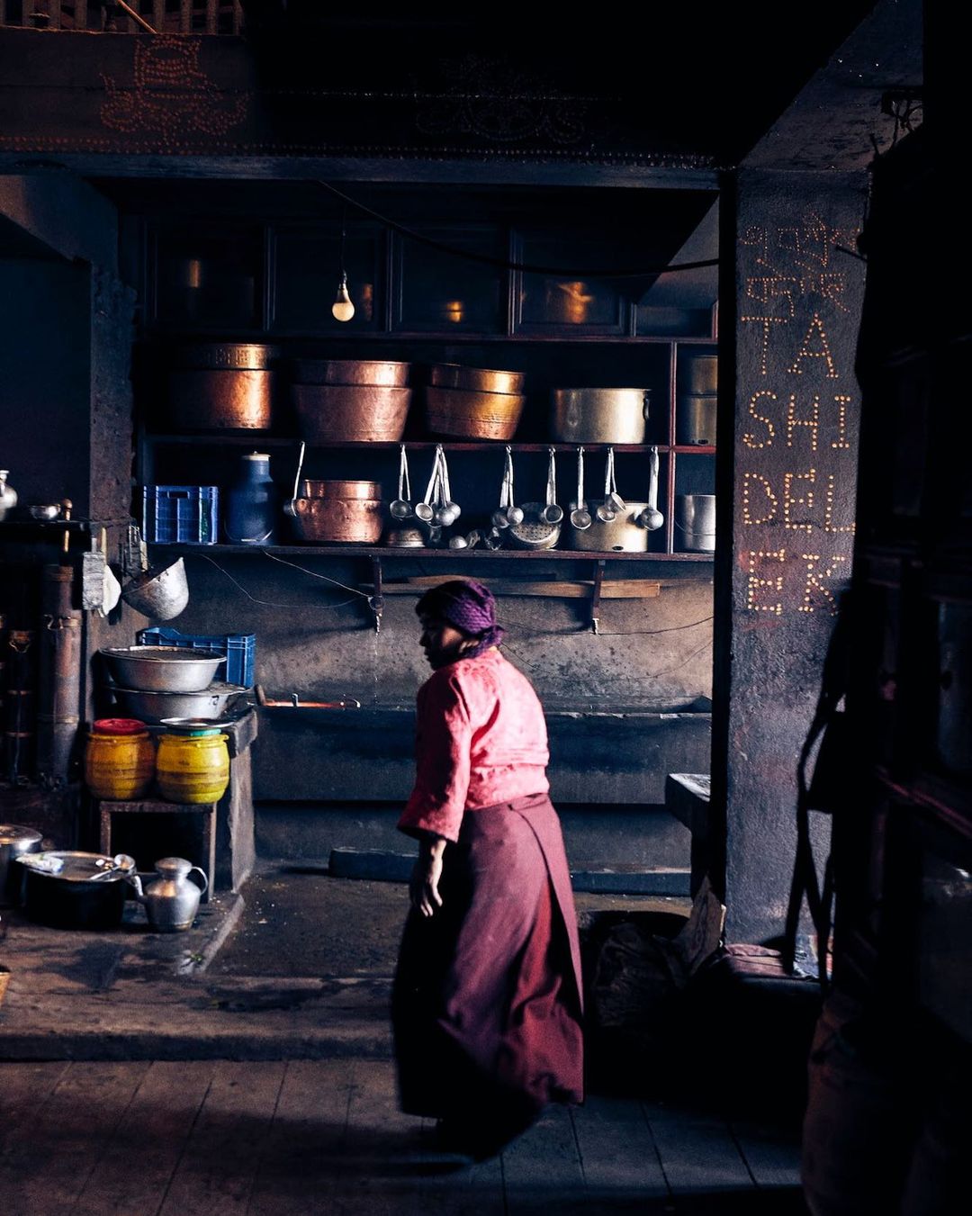 Woman walks through a rustic kitchen with copper pots hanging on shelves, captured in a dimly lit, vintage style interior scene.