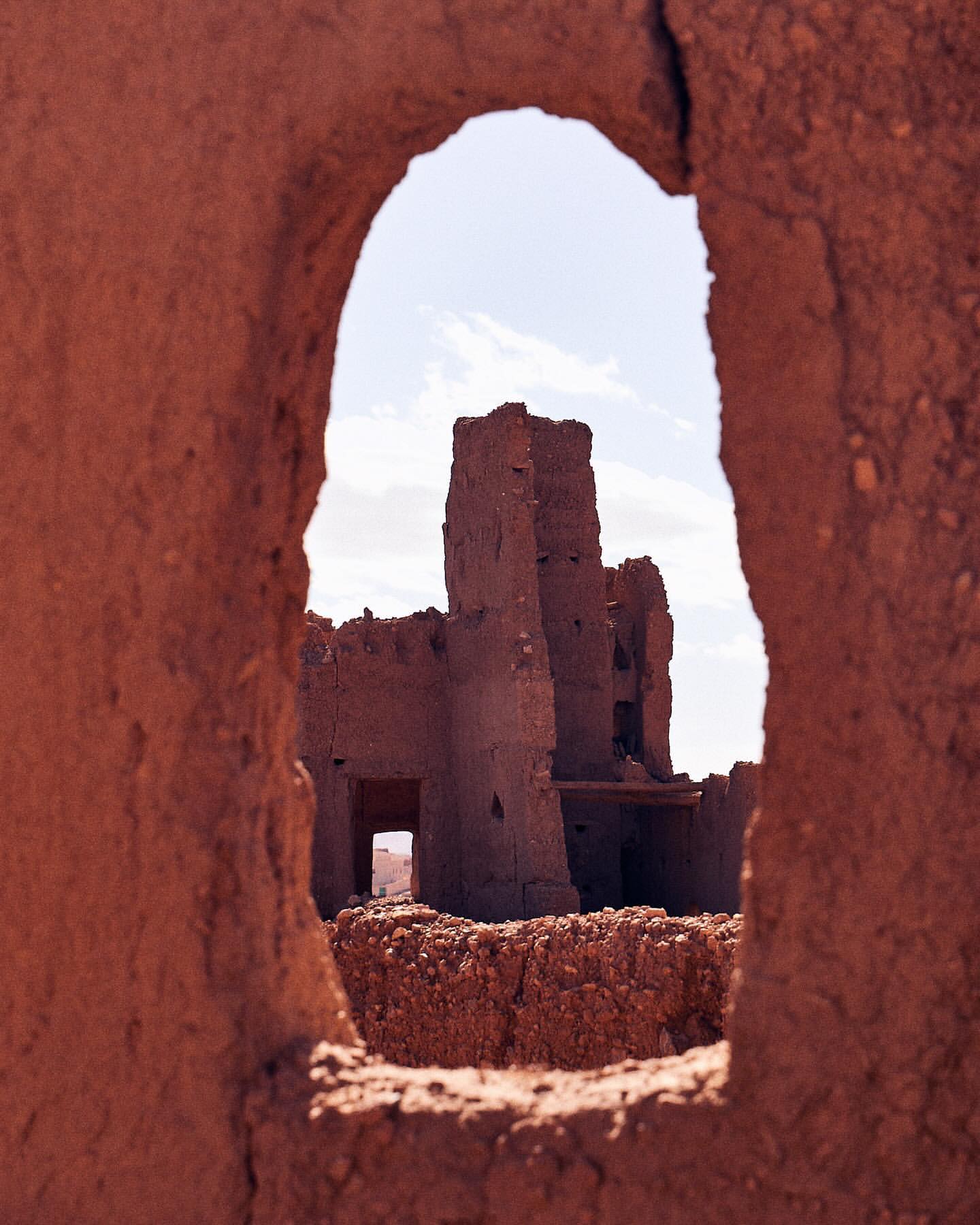 Ruined adobe building seen through an eroded archway in a desert landscape under a bright, slightly cloudy sky.