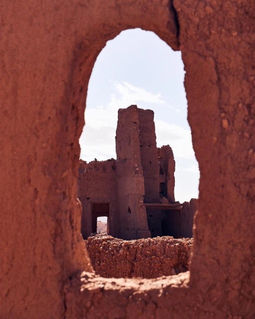 Ruined adobe building seen through an eroded archway in a desert landscape under a bright, slightly cloudy sky.