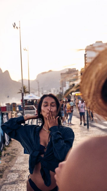 Woman in bikini and unbuttoned shirt poses blowing a kiss on a cobblestone path with people and mountains in the background.