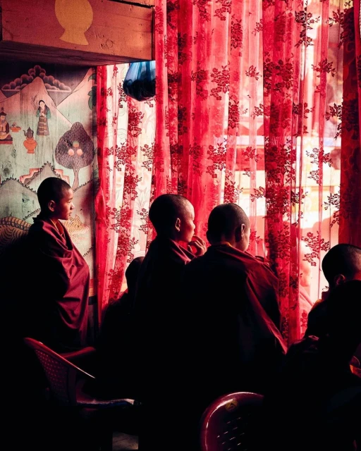 Young monks in maroon robes are seated in a dimly lit room, set against a hand-painted wall and red floral-patterned curtains.