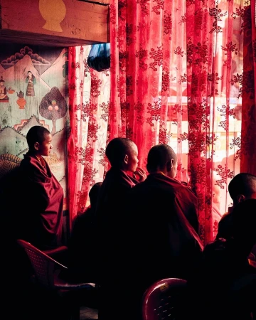 Young monks in maroon robes are seated in a dimly lit room, set against a hand-painted wall and red floral-patterned curtains.