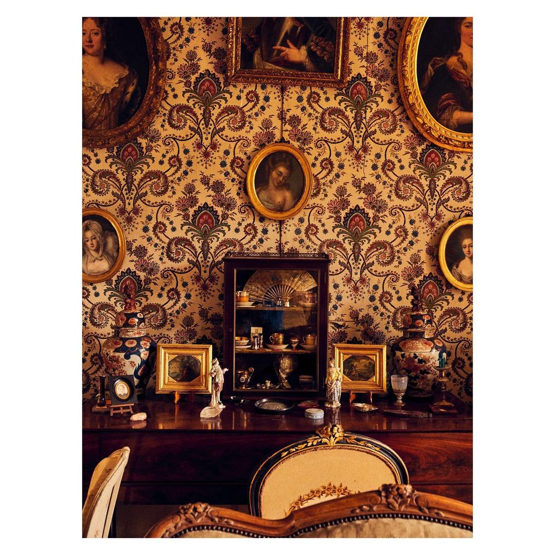 Vintage interior with ornate wallpaper, framed portraits, display cabinet, and decorative objects on a wooden dresser with chair detail.