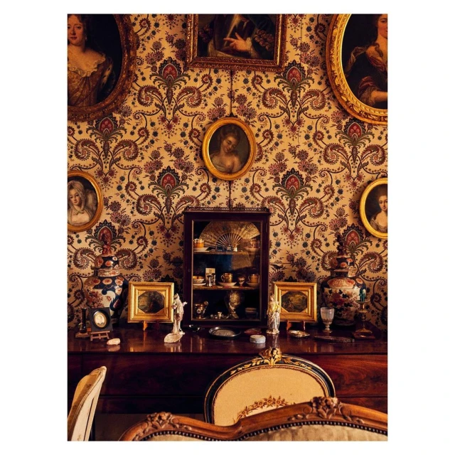 Vintage interior with ornate wallpaper, framed portraits, display cabinet, and decorative objects on a wooden dresser with chair detail.