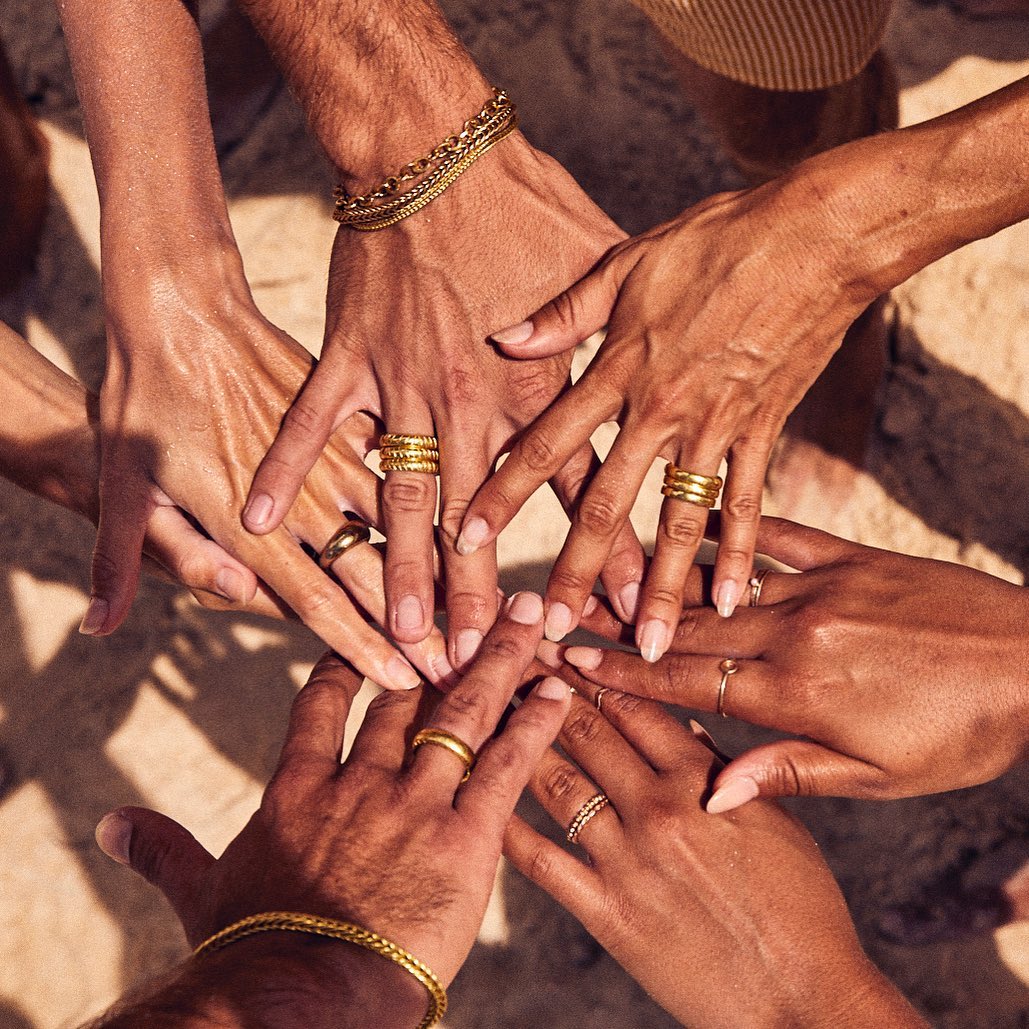 A group of people joins hands, showcasing various gold rings and bracelets, with a light-brown, textured backdrop adding warmth to the image.