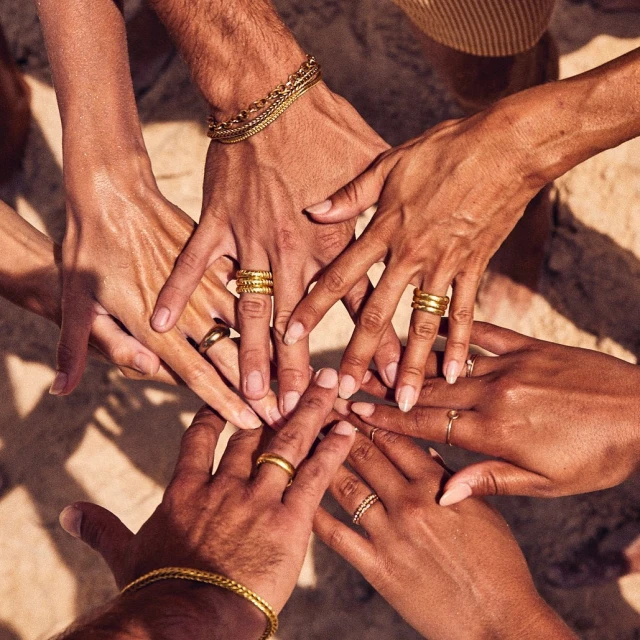 A group of people joins hands, showcasing various gold rings and bracelets, with a light-brown, textured backdrop adding warmth to the image.