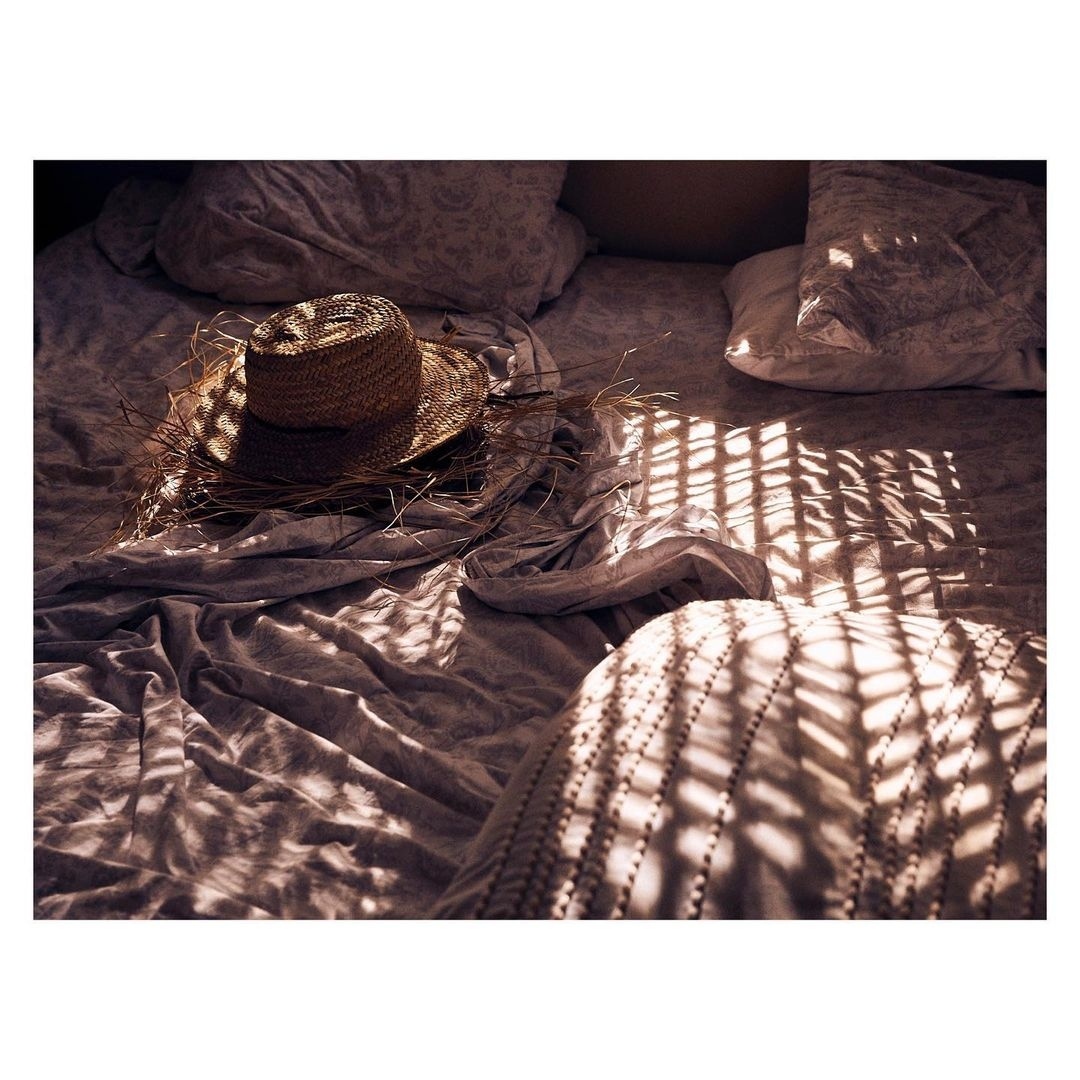 Straw hat rests on a rumpled bed with morning sun casting patterned shadows across the bedding and a knitted pillow in the scene.