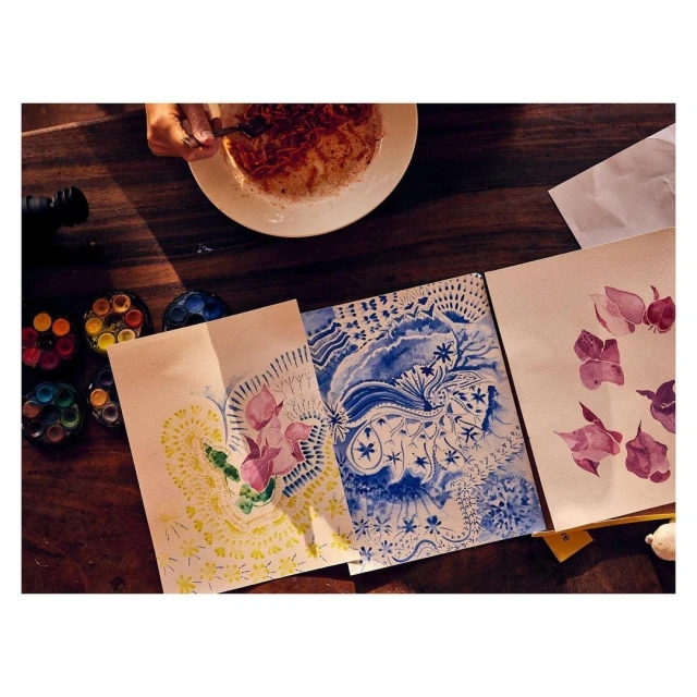 Overhead shot of watercolor paintings of flowers on a dark wooden table with art supplies and a person eating in the background.