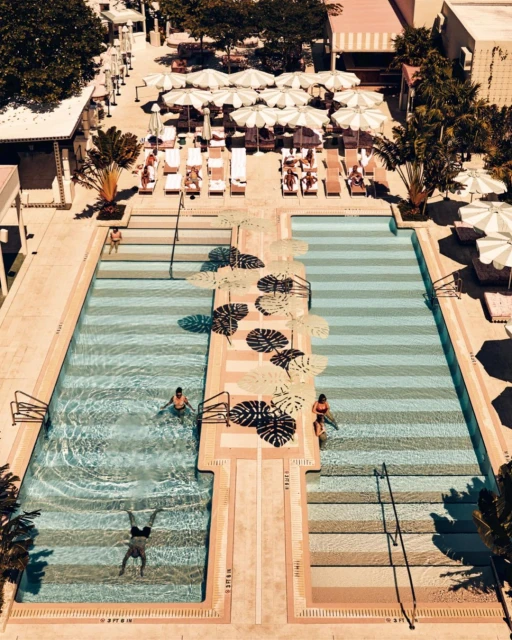 Aerial view of a luxury hotel swimming pool area with palm trees, lounge chairs, umbrellas, and people swimming and sunbathing outdoors.
