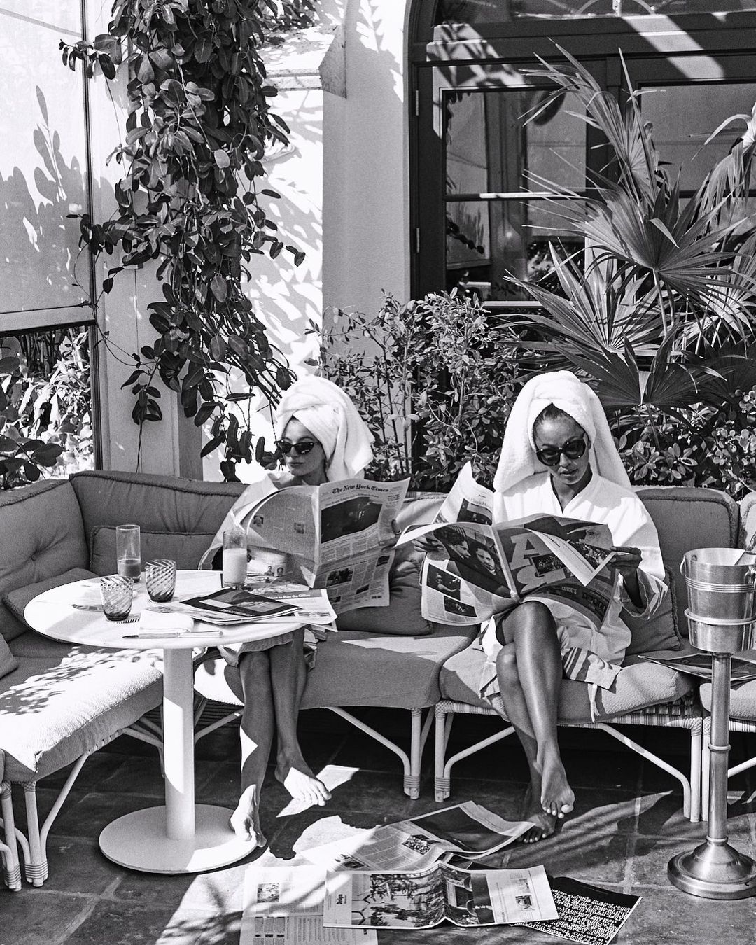 Two women in robes and towels read newspapers on a patio. Black and white photo shows them at a table with drinks outside.