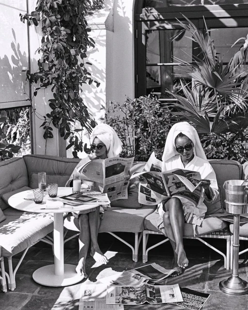 Two women in robes and towels read newspapers on a patio. Black and white photo shows them at a table with drinks outside.