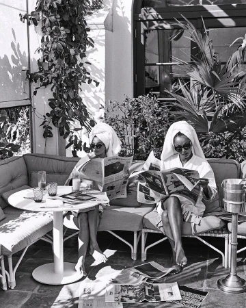 Two women in robes and towels read newspapers on a patio. Black and white photo shows them at a table with drinks outside.