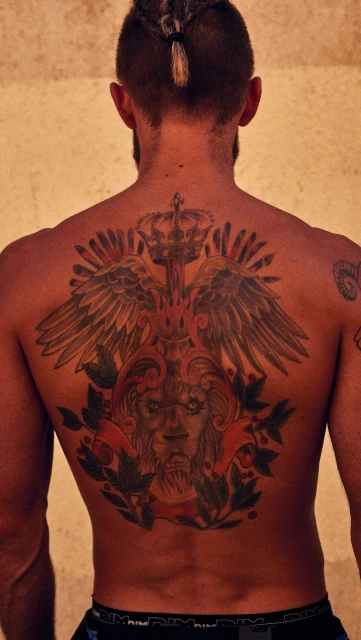 Back view of a man with a tattoo of a lion and eagle on his upper back, showing his skin texture and braided hair detail.