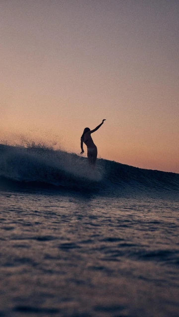 Female surfer riding a wave at sunset with a pink sky, creating a silhouette on the ocean water.