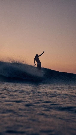 Female surfer riding a wave at sunset with a pink sky, creating a silhouette on the ocean water.