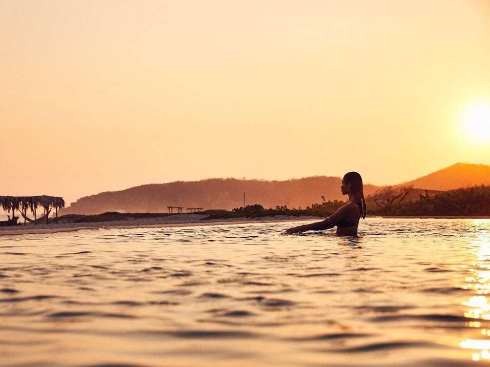 Woman in ocean at sunrise. Backlit silhouette of adult female wading in water with mountains and beach in background with orange sky.