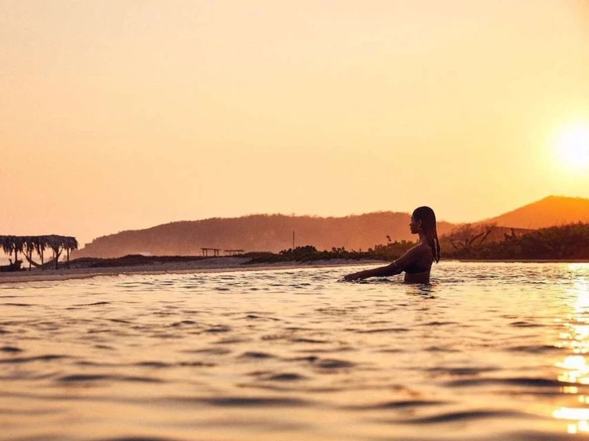 Woman in ocean at sunrise. Backlit silhouette of adult female wading in water with mountains and beach in background with orange sky.
