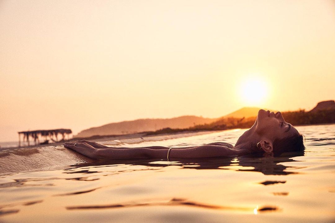 Person lying on their back in water at the beach during golden hour, with a building and hills visible in the distance.