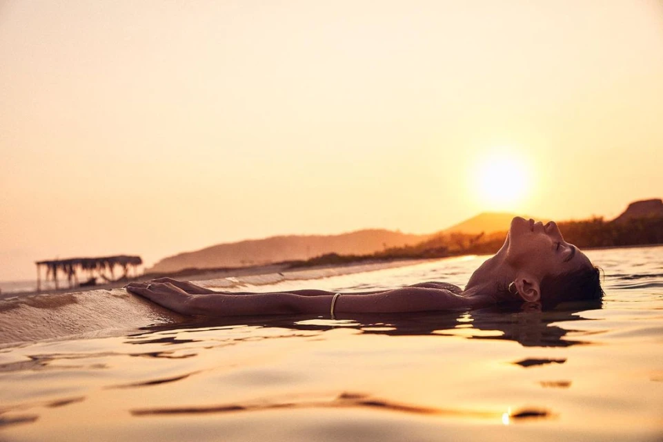 Person lying on their back in water at the beach during golden hour, with a building and hills visible in the distance.