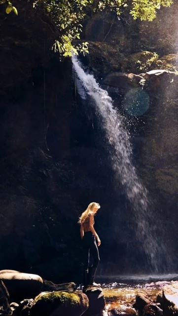 A woman stands near the base of a cascading waterfall, surrounded by lush greenery, sunlight and moss-covered rocks in a nature scene.