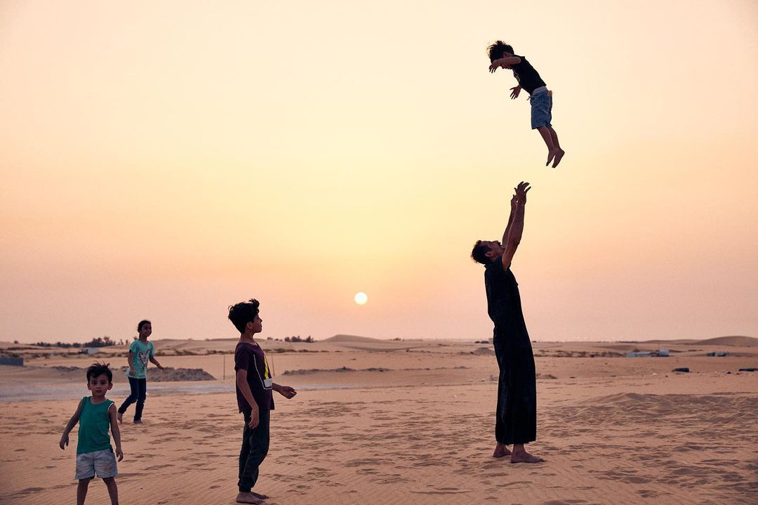Adult man throwing a child into the air on a sandy desert landscape at sunset with a group of other children nearby, creating a joyful scene.
