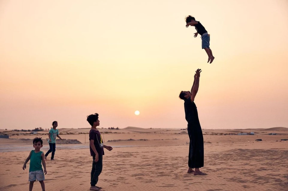 Adult man throwing a child into the air on a sandy desert landscape at sunset with a group of other children nearby, creating a joyful scene.