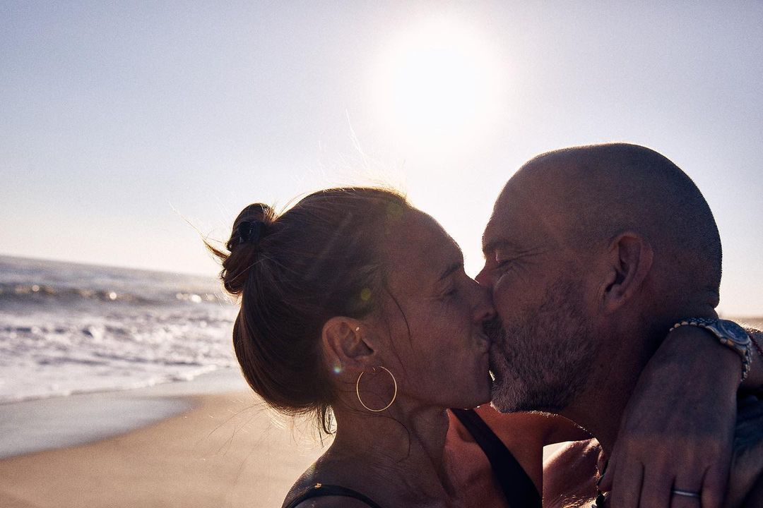 Silhouetted adult couple kisses on a beach, sun shining behind them with ocean waves in the background on a sunny day. Lifestyle shot.