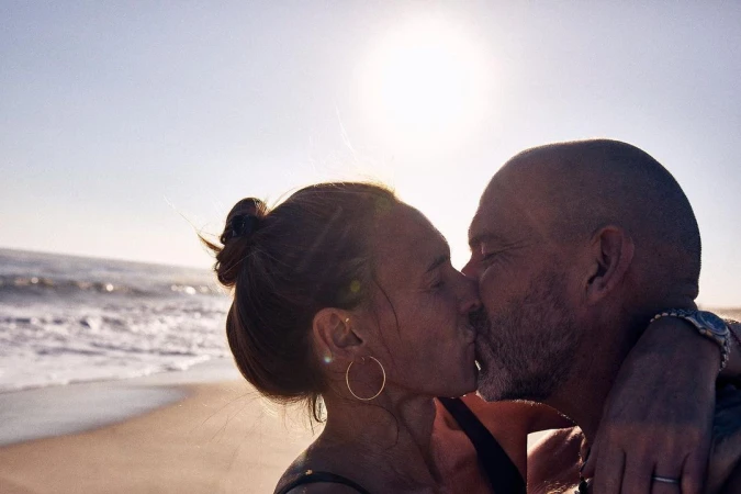 Silhouetted adult couple kisses on a beach, sun shining behind them with ocean waves in the background on a sunny day. Lifestyle shot.