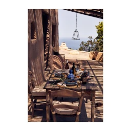 Wooden dining table set on a sunlit outdoor terrace with wine bottles, glassware, and dishes under a pergola.