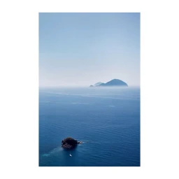 Aerial seascape photography by Oliver Pilcher showing a small rocky island and boat in blue water with distant islands in haze under a clear sky