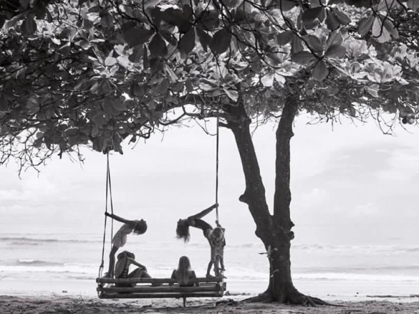 Black and white photograph of people on a large wooden swing hanging from a leafy tree branch on a sandy beach.