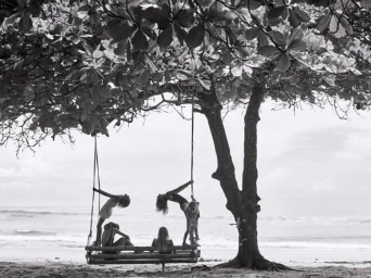 Black and white photograph of people on a large wooden swing hanging from a leafy tree branch on a sandy beach.