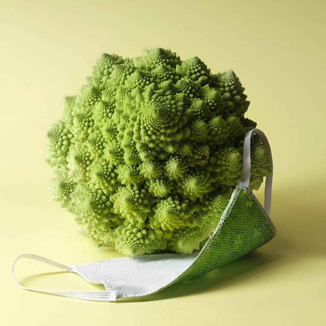 Still life studio shot of a romanesco broccoli and a fabric face mask on a pale yellow background.