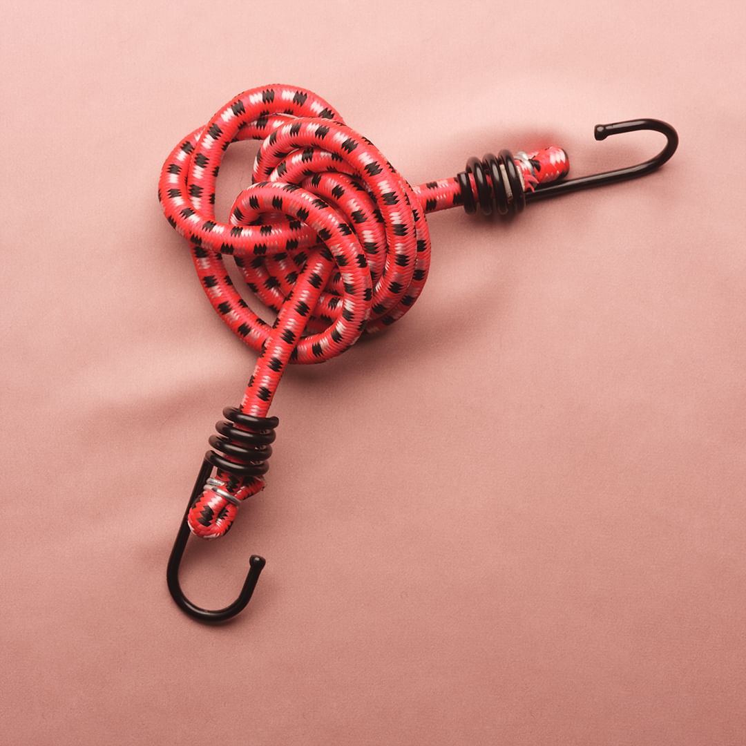 Overhead shot of a red and black bungee cord with metal hooks, coiled on a tan surface, product, gear, equipment.