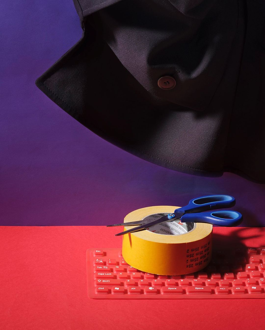 Studio shot of scissors cutting tape roll atop red keyboard against purple backdrop, topped by partially visible dark garment.