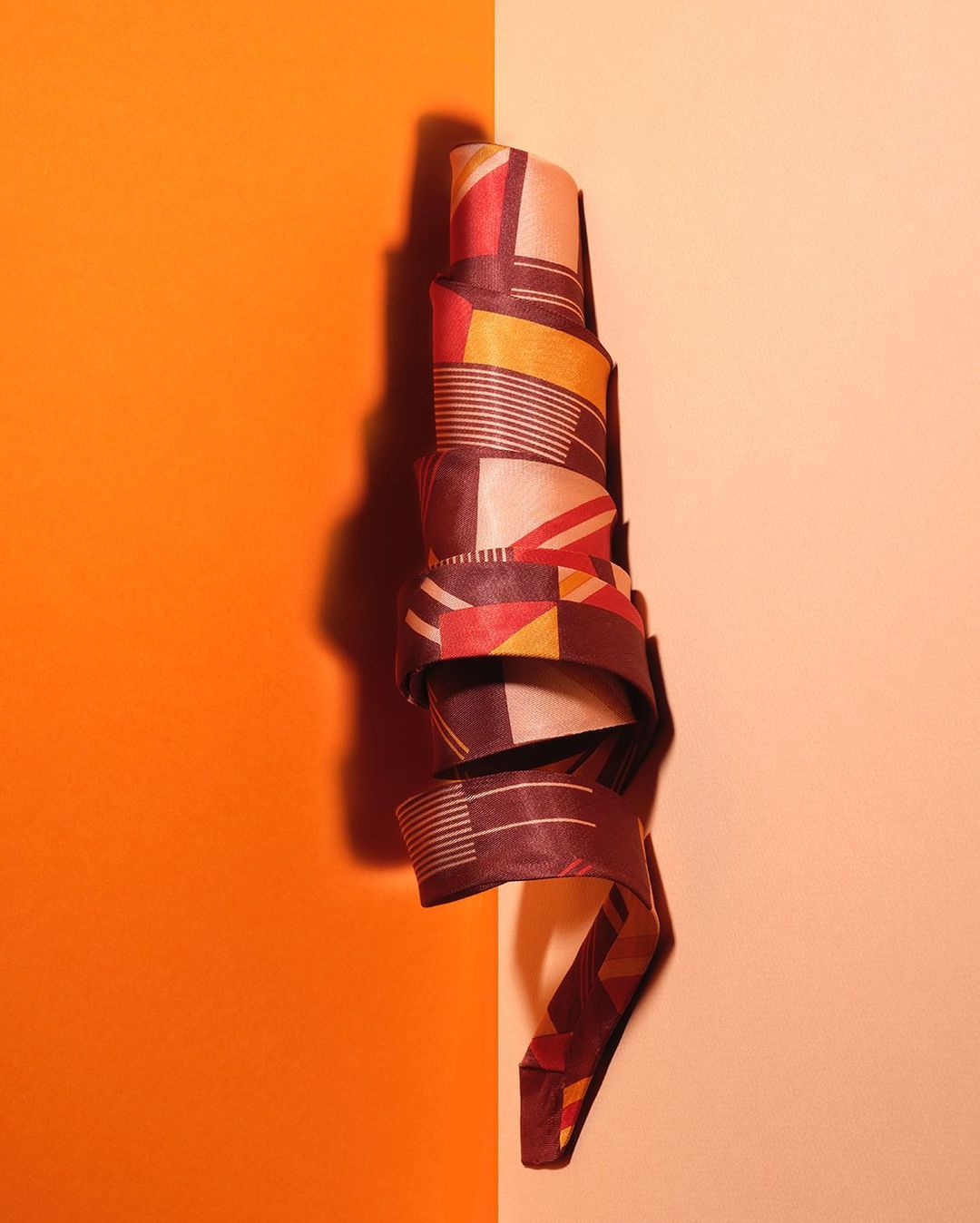 Abstract geometric pattern featuring brown, red, yellow, and white on a curled paper strip against orange and peach backgrounds.
