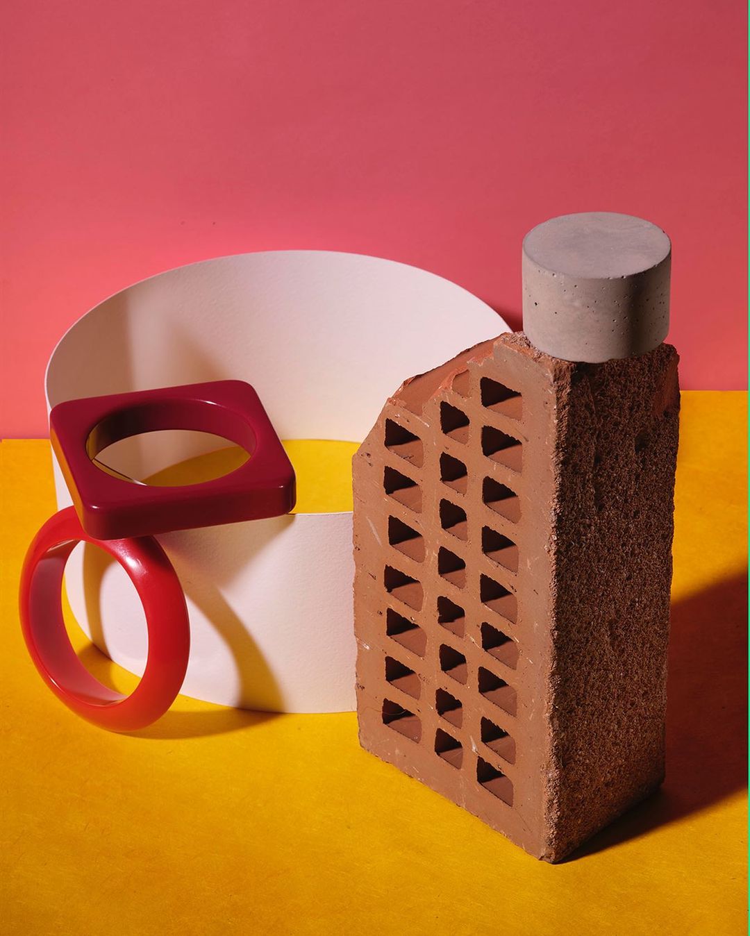 Still life composition with a broken red brick, concrete cylinder, geometric red bracelets, and white shapes against a pink and yellow background.