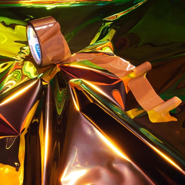 Shiny iridescent material is partially covered with copper-toned tape, partially unrolled to reveal the shiny yellow material underneath.